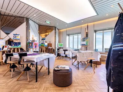 Ein helles Atelier mit Holzboden und Meerblick an Bord der Mein Schiff 1. Mehrere Staffeleien mit bunten Bildern, Tische mit weissen Tischtüchern und Stühle sind zu sehen. Ein Schiffs-Motiv auf einer Staffelei.