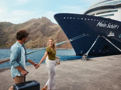 Ein Paar mit Gepäck geht Hand in Hand auf einem sonnigen Pier. Im Hintergrund ist die Mein Schiff 1 vertäut und eine malerische Berglandschaft zu sehen.