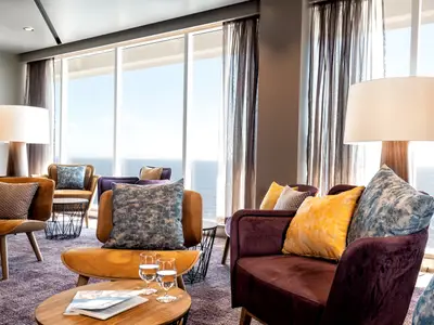 Helle, moderne Lounge an Bord der Mein Schiff 1 mit Panoramafenstern zum Meer. Gemütliche Sessel in Gelb und Lila mit Kissen. Ein grosser Lampenschirm und Gläser auf einem Tisch sind zu sehen.