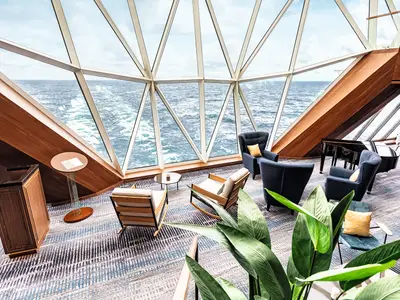 Eine moderne Lounge auf der Mein Schiff 1 mit grossen, geometrischen Fenstern, die den Blick auf das weite Meer freigeben. Mehrere Sessel und ein Flügel laden zum Verweilen ein.