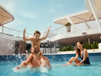 Familie hat Spaß im Pool der Mein Schiff 1. Vater trägt Sohn auf Schultern, spritzt Wasser. Mutter sitzt lächelnd daneben. Sonnige, lebhafte Szene auf dem Deck.