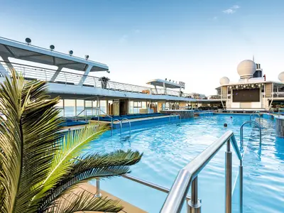 Weitwinkelaufnahme des großen Außenpools auf dem Oberdeck der Mein Schiff 1, umgeben von Sonnenliegen, Palmen und einem oberen Deck mit Meerblick. Mehrere Personen schwimmen im Pool.