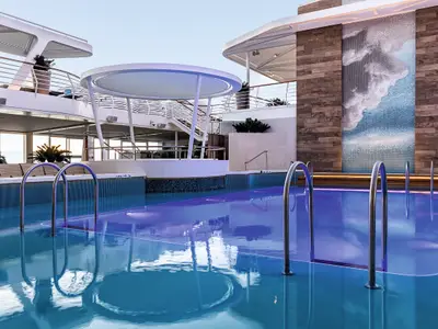 The 'Lagune' pool area on Mein Schiff 1 at dusk. The blue-purple illuminated pool features metal ladders, surrounded by stylish wooden walls, a cloud mosaic, and multi-level decks with lounge seating.