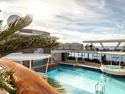 Sonnige Pool-Lagune der Mein Schiff 1. Großer Pool, Whirlpool und viele Liegestühle. Meer im Hintergrund, Palmen im Vordergrund. Wenige Gäste entspannen sich.