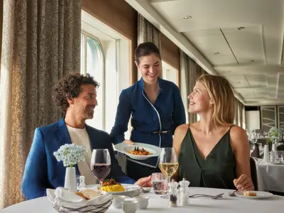 Ein Paar wird in einem Restaurant an Bord der Mein Schiff 1 von einer Crew-Mitarbeiterin bedient. Sie serviert ein Gericht an den gedeckten Tisch mit Wein und Blumen.