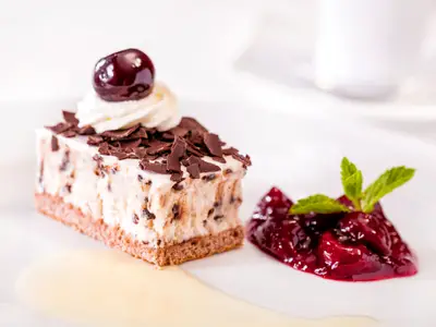 Ein Stück Schwarzwälder Kirschtorte mit Sahne, Schokoraspeln und einer Kirsche obendrauf, serviert auf einem weißen Teller mit Kirschsauce und Minze daneben an Bord der Mein Schiff 1.