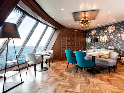 Ein helles Restaurant an Bord der Mein Schiff 1 mit grossen Fenstern zum Meer. Blaue Stühle, Holztische und eine Sitzbank mit floraler Tapete schmücken den Raum.
