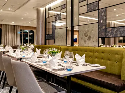 Ein stilvolles Restaurant an Bord der Mein Schiff 1 mit mehreren elegant gedeckten Tischen und bequemen olivgrünen Polsterbänken. Im Hintergrund sind große Fenster und eine dekorative Wandverkleidung aus Spiegeln und strukturierten Platten zu sehen. Jeder Tisch ist mit weißen Stoffservietten, Besteck, Gläsern und blauen Trinkgläsern versehen. Blumenarrangements schmücken einige Tische.