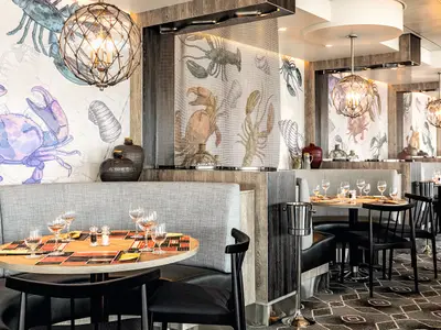 Blick in das Fischmarkt-Restaurant an Bord der Mein Schiff 1. Mehrere Tische mit Stühlen und gepolsterten Bänken sind elegant gedeckt. Die Wände zieren großflächige Zeichnungen von Meeresfrüchten wie Krebsen und Hummern. Elegante Pendelleuchten tauchen den Raum in warmes Licht.