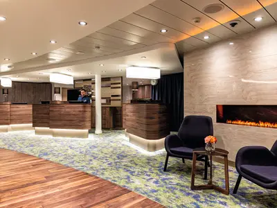 Der stilvolle Rezeptionsbereich der Mein Schiff 1. Dunkelbraune Holztheken, ein Mitarbeiter, moderne Teppichböden und eine gemütliche Kaminlounge mit Sesseln sind zu sehen.