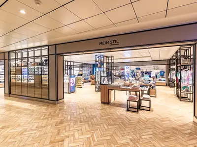 Blick in den hell beleuchteten "Mein Stil by Heinemann" Shop an Bord der Mein Schiff 1 mit Regalen voller Mode, Taschen und Accessoires.