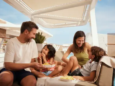 Eine lächelnde Familie isst Pommes an Deck der Mein Schiff 1. Der Vater reicht der Tochter eine Schale. Im Hintergrund ist ein Poolbereich unter blauem Himmel zu sehen.