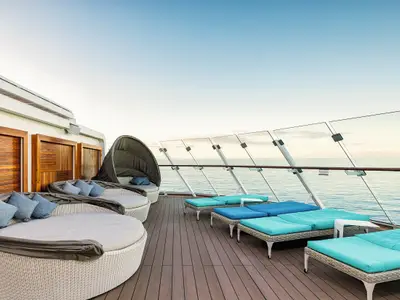 Sonnendeck der Mein Schiff 1 mit Daybeds und türkisen Liegen auf Holzdeck. Glasbalustrade zum Meer unter blauem Himmel. Eine entspannte Umgebung.