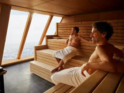 Zwei Männer in weißen Handtüchern entspannen in einer modernen Holzsauna mit großen Panoramafenstern, die den Blick auf das weite Meer an Bord der Mein Schiff 1 freigeben.