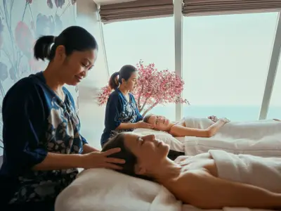 Zwei Frauen liegen auf Massageliegen im Spa der Mein Schiff 1 und erhalten Kopfmassagen von Therapeutinnen. Im Hintergrund ist ein großer Fensterbereich mit Meerblick zu sehen, davor steht ein blühender Baum.