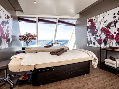 Ein luxuriöser Spa-Anwendungsraum an Bord der Mein Schiff 1 mit zwei Massageliegen und grossen Fenstern, die einen weiten Blick auf das offene Meer bieten. Die Wände sind mit floralen Kunstwerken verziert.