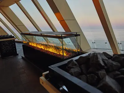 Die moderne finnische Sauna an Bord der Mein Schiff 1. Zu sehen sind zwei Saunaöfen mit Steinen und einen langen Elektrokamin. Grosse Fenster mit Meerblick bei Sonnenuntergang.