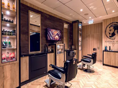 Innenansicht des Barbershop Hairspa an Bord der Mein Schiff 1 mit zwei schwarzen Friseurstühlen, Holzvertäfelung und Regalen voller Pflegeprodukte. Ein Fernsehbildschirm zeigt Rasierutensilien.