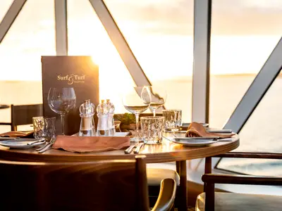 Ein elegant eingedeckter Tisch für zwei Personen im Surf & Turf Steakhouse an Bord der Mein Schiff 1. Im Hintergrund ist der Ozean bei Sonnenuntergang zu sehen.