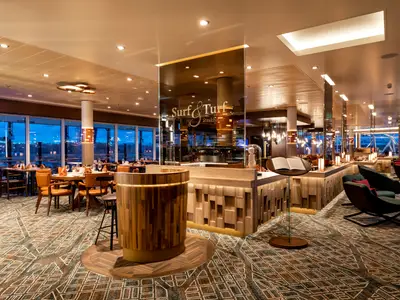 Das Surf & Turf Steakhouse an Bord der Mein Schiff 1 bei blauer Stunde. Elegante Tische mit Holzstühlen und großen Panoramafenstern dominieren den Raum. Eine Holztheke und Lichtinstallationen sorgen für eine warme Atmosphäre.
