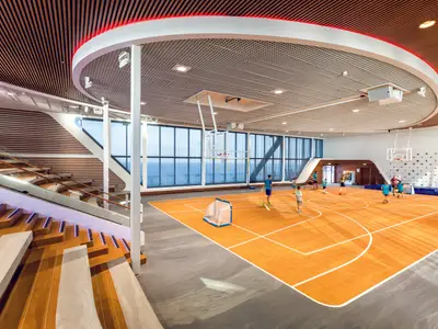 Blick in die helle Arena an Bord der Mein Schiff 1, wo Kinder auf einem orangen Sportfeld spielen. Große Fensterfronten bieten Meerblick, links eine Treppe, rechts eine Kletterwand.