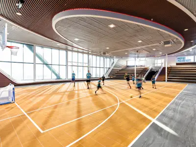 Die helle, moderne Arena an Bord der Mein Schiff 1 zeigt eine Gruppe von Personen, die auf einem Holzboden spielen. Große Fensterfronten bieten viel Tageslicht. Ein Basketballkorb und ein kleines Fußballtor sind sichtbar. Einige Personen sind durch Bewegung leicht unscharf.