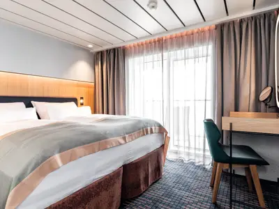 Blick in eine helle Suite an Bord der Mein Schiff 1. Komfortables Doppelbett, Schminktisch mit Spiegel und Fensterfront mit Gardinen, die Tageslicht spendet.
