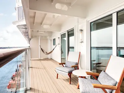Geräumiger Balkon einer Suite an Bord der Mein Schiff 1 mit modernem Holzboden, eleganten Sesseln und einer Hängematte. Im Hintergrund ist das Meer und ein Hafen zu sehen.