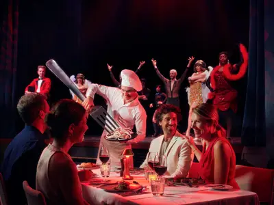 An Bord der Mein Schiff 1 genießen Gäste ein lebhaftes Varieté Dinner. Ein Koch serviert riesige Spaghetti, während Tänzer auf der Bühne auftreten. Eine festliche Abendstimmung.