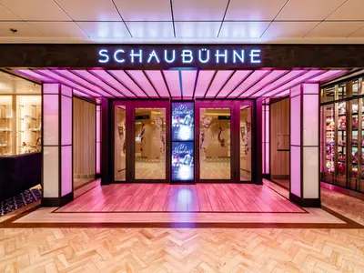 Eingang zur Schaubühne an Bord der Mein Schiff 1. Rosa Beleuchtung akzentuiert den Bereich. Über den Türen leuchtet 'SCHAUBÜHNE'. Bildschirme zeigen 'It's Showtime'.