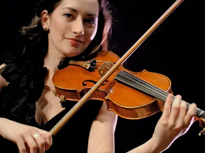 Nahaufnahme einer jungen Frau mit dunklen Haaren, die eine Violine spielt und lächelt. Sie trägt ein schwarzes, gerafftes Oberteil und Ohrringe. Die Szene ist an Bord der Mein Schiff 2 bei schlechten Lichtverhältnissen. Ihre rechte Hand hält den Bogen über die Saiten, während ihre linke Hand die Saiten am Griffbrett drückt.