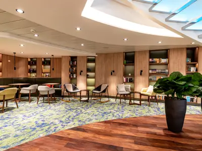 Ein breiter Blick auf eine gemütliche Lounge an Bord der Mein Schiff 2. Links gibt es eine durchgehende dunkelgrüne Lederbank mit Holztischen. Davor stehen hellgraue Stühle und ein senfgelber Sessel auf einem blau-grün gemusterten Teppich. Rechts sind Bücherregale in eine Holzvertäfelung integriert, davor stehen Sessel und zwei große Topfpflanzen. Das Licht kommt von Deckenleuchten und beleuchteten Regalen.