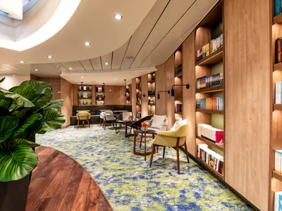 Ein einladender Bereich an Bord der Mein Schiff 2 mit einer Vielzahl von Büchern in beleuchteten Holzregalen. Es gibt mehrere moderne Sessel und kleine Tische auf einem Teppichboden mit grünen und blauen Mustern. Im Vordergrund befindet sich eine große Pflanze mit grünen Blättern.