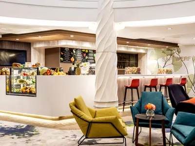 Blick in die moderne, helle Saftwerft der Mein Schiff 2 mit einer langen Bar, an der frische Früchte ausgestellt sind. Gegenüber stehen gemütliche Sessel in gelb, türkis und dunkelblau.