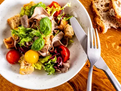 Ein appetitlicher Salat mit zartem Fleisch, knusprigen Croutons, frischen Tomaten und Kräutern, serviert auf einem weißen Teller an Bord der Mein Schiff 2. Daneben liegen Besteck und angeschnittenes Brot auf einem Holztisch.