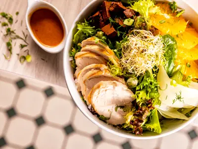 Ein appetitlicher Hähnchensalat in einer weißen Schüssel, dazu eine kleine Schale mit Dressing. Der Salatteller, wie er auch an Bord der Mein Schiff 2 serviert werden könnte, ist reich an frischem Grün, Sprossen, Orangen und geschnittenem Hähnchen. Eine gesunde Mahlzeit auf einem hellen Holztisch, im Hintergrund ist ein gemusterter Boden erkennbar.