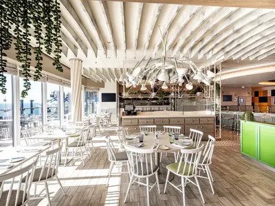 Innenansicht eines hellen Restaurants auf der Mein Schiff 2. Weiße Tische und Stühle stehen vor großen Fenstern mit Blick auf das Meer. Über den Tischen hängt eine moderne weiße Kronleuchter-Installation. Im Hintergrund sind weitere Essbereiche und eine Theke zu sehen.