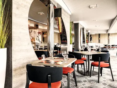 Ein helles und modern eingerichtetes Restaurant an Bord der Mein Schiff 2. Mehrere Tische mit dunklen Stühlen und roten Sitzkissen sind gedeckt mit Gläsern. Die Wände sind teils holzvertäfelt, teils mit maritimen Motiven bemalt. Dekorative Pflanzen und Spiegelsäulen zieren den Raum.