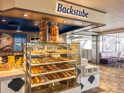 Innenansicht der Bäckerei "Backstube" an Bord der Mein Schiff 2. Eine Glasvitrine zeigt zahlreiche süße und herzhafte Backwaren. Links daneben sind glutenfreie Produkte auf einer Theke. Im Hintergrund ist eine gemütliche Sitzecke mit lilafarbenen Bänken und Tischen.