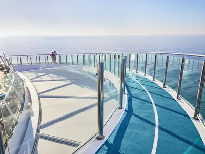 Eine erhöhte Perspektive auf ein offenes Deck der Mein Schiff 2. Links sind Tische und Stühle eines Außenbereichs zu sehen. In der Mitte des Bildes sind zwei Personen auf einem Laufweg unterwegs. Rechts befindet sich eine blaue Laufbahn, die sich um eine Glasbrüstung herum schlängelt. Im Hintergrund erstreckt sich das weite, blaue Meer bis zum Horizont unter einem klaren Himmel. Die Sonne spiegelt sich auf der Wasseroberfläche.