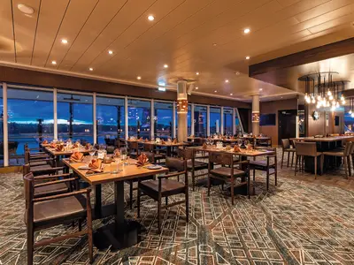 Ein geräumiges Restaurant an Bord der Mein Schiff 2 bei Dämmerung, mit Holztischen, braunen Stühlen und großen Panoramafenstern. Draußen ist das Wasser und der Horizont zu sehen. Die Decke ist mit Einbaustrahlern beleuchtet, und im hinteren Bereich hängen auffällige Leuchter über einer langen Theke.