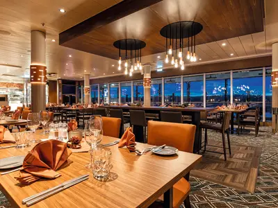 Ein elegantes Restaurant auf der Mein Schiff 2 mit Tischen, Stühlen und einer langen Bar. Große Fenster bieten einen Panoramablick auf die Stadt bei Einbruch der Dunkelheit. Die Tische sind mit orangefarbenen Servietten und Gläsern gedeckt.