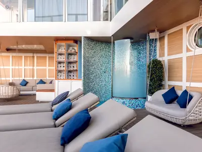 Blick auf eine luxuriöse Outdoor-Lounge an Bord der Mein Schiff 2 mit grauen Liegestühlen, blauen Kissen und zwei hängenden Korbsesseln. Im Hintergrund befindet sich eine Duschkabine mit türkisfarbenen Mosaikfliesen und ein offenes Regal mit gerollten Handtüchern.
