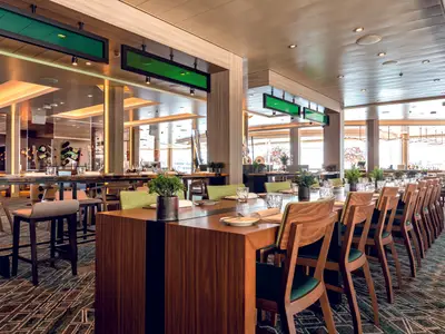Blick in ein geräumiges, helles Restaurant an Bord der Mein Schiff 2. Lange Holztische mit grünen Stühlen und Topfpflanzen sind festlich gedeckt. Im Hintergrund sind große Fenster und eine Bar zu sehen.