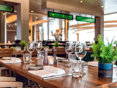 Ein Detailblick auf einen gedeckten Tisch im Restaurant des Kreuzfahrtschiffs Mein Schiff 2. Der Holztisch ist mit Gläsern, Tellern, Besteck, Servietten, einer Kerze, Pfeffer- und Salzmühlen und einer kleinen grünen Pflanze dekoriert. Im Hintergrund sind weitere Tische und der Innenbereich des Restaurants mit einem Fenster zu sehen.