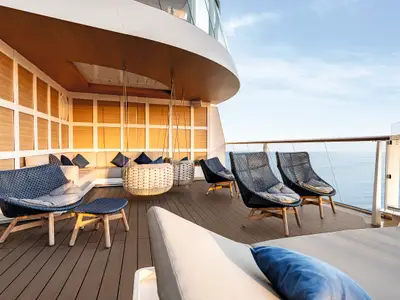Sonnendeck der Mein Schiff 2 mit Sitzgelegenheiten aus Rattan und Holz, darunter eine L-förmige Couch, zwei Hängesessel, drei Sessel und eine Liege. Das Meer ist im Hintergrund zu sehen. Ein Glasgeländer trennt das Deck vom Meer.