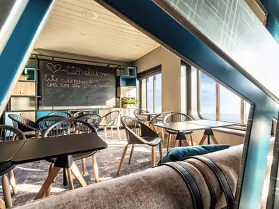 Innenansicht eines Aufenthaltsraumes der Mein Schiff 2 mit dunklen Tischen und Stühlen, einer großen Tafel mit einer Willkommensnachricht und Fenstern, die einen weiten Blick auf das offene Meer bieten. Der Raum ist in Blautönen gehalten.