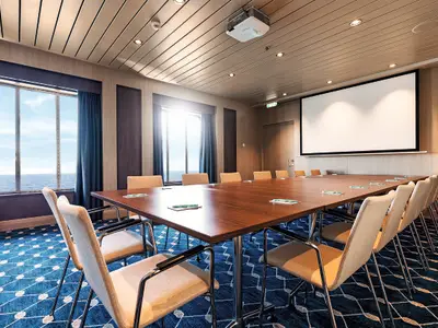 Ein Konferenzraum auf der Mein Schiff 2 mit einem langen Holztisch, bequemen Stühlen, einem großen Fenster mit Meerblick und einem ausfahrbaren Projektionsbildschirm an der Wand.