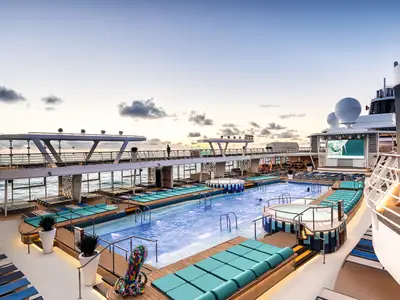 Dieses Pressefoto zeigt das weitläufige Pooldeck der Mein Schiff 2 in der Abenddämmerung. Zwei große Pools, umgeben von türkisfarbenen Liegen und Holzdecks, erstrecken sich unter einem Brückenbereich. Im Hintergrund ist das offene Meer zu sehen und eine große Leinwand zeigt ein Bild. Ein paar Menschen sind in den Pools. Das Schiff ist hell beleuchtet.