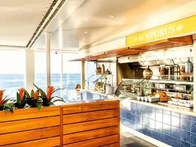 Blick auf das türkische Restaurant 'Bosphorus Fast & Slow Food' an Bord der Mein Schiff 2. Links große Fenster mit Meerblick, rechts ein Buffet mit türkischen Spezialitäten wie Döner. Dekorative Elemente mit Holz und blauen Fliesen sind zu sehen.